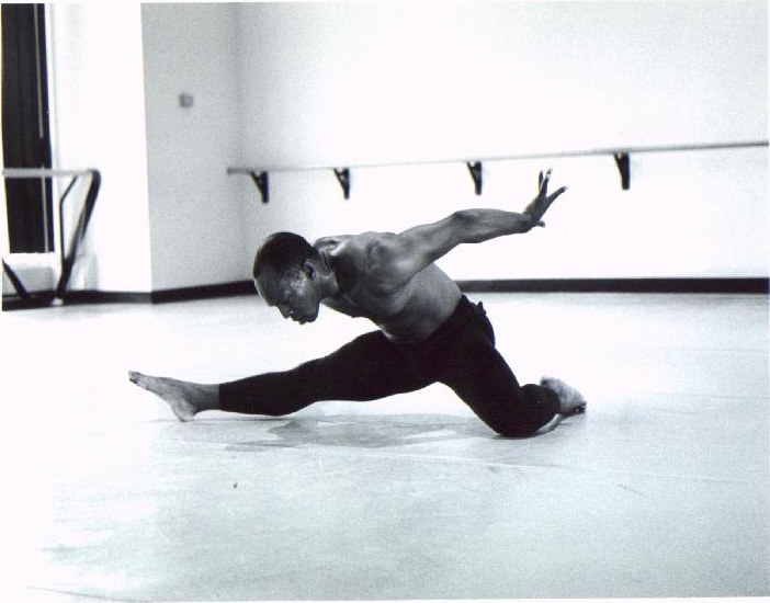 Black-and-white photograph of Boris Willis in a low lunge along the studio floor, one arm reaching forward, beside the ballet barre
