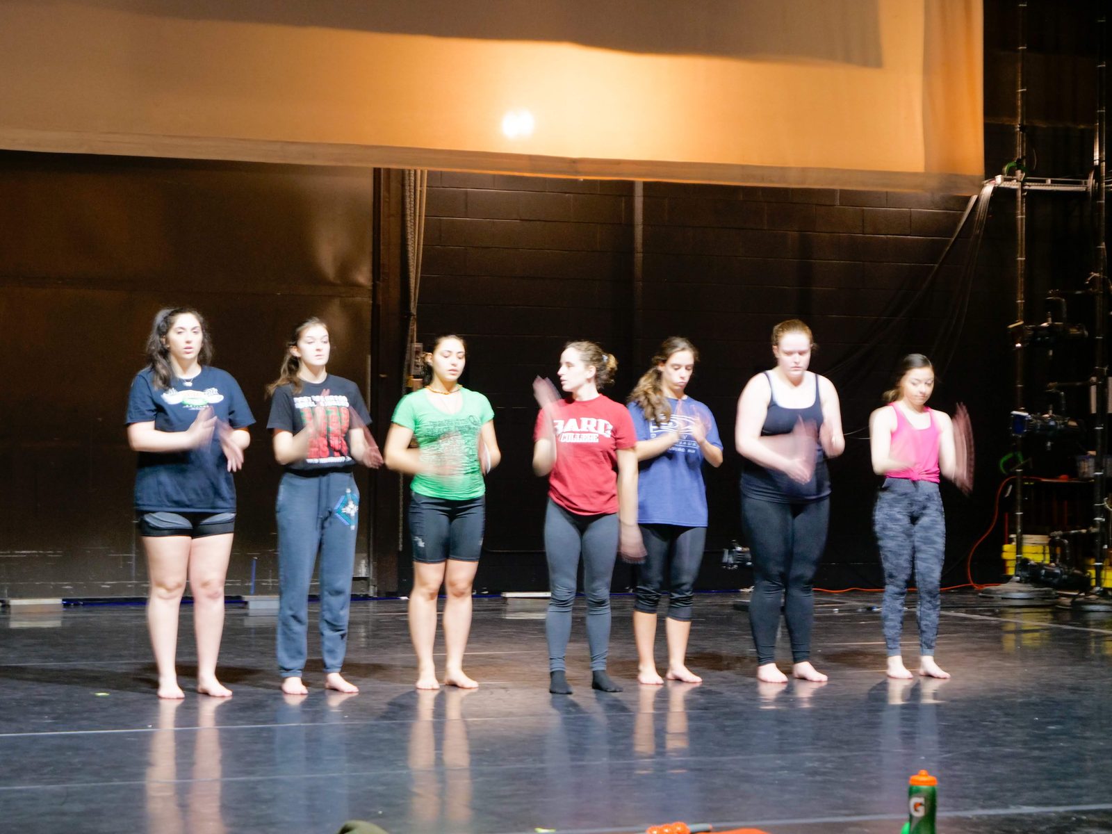 A line of seven dancers standing across the stage in rehearsal clothes, work lights up, theater fly system visible at right