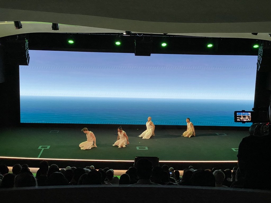 Four dancers kneeling before an open-ocean horizon rendered in Unreal Engine