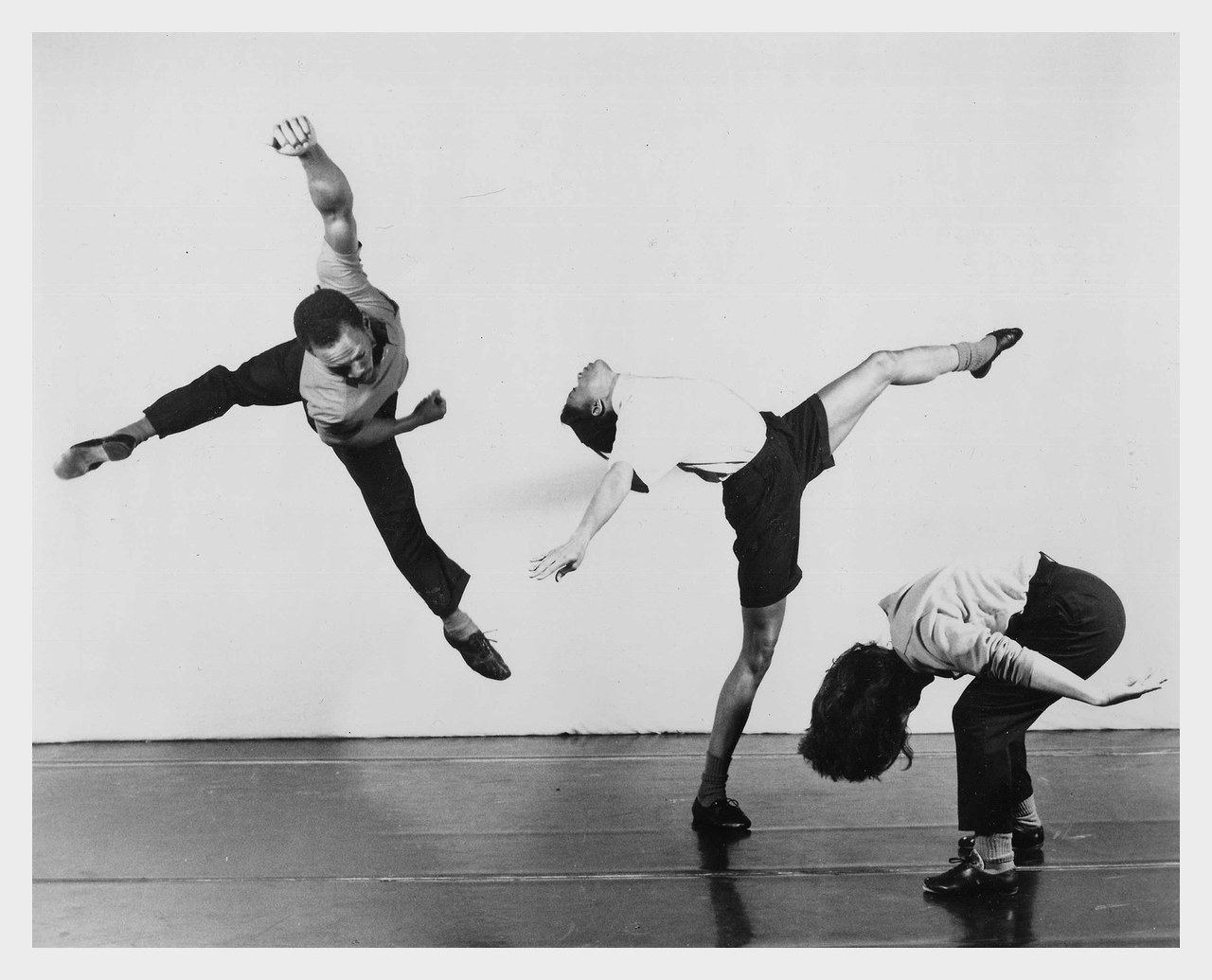 Black-and-white performance photograph of three dancers in mid-movement, one leaping with arms flung wide