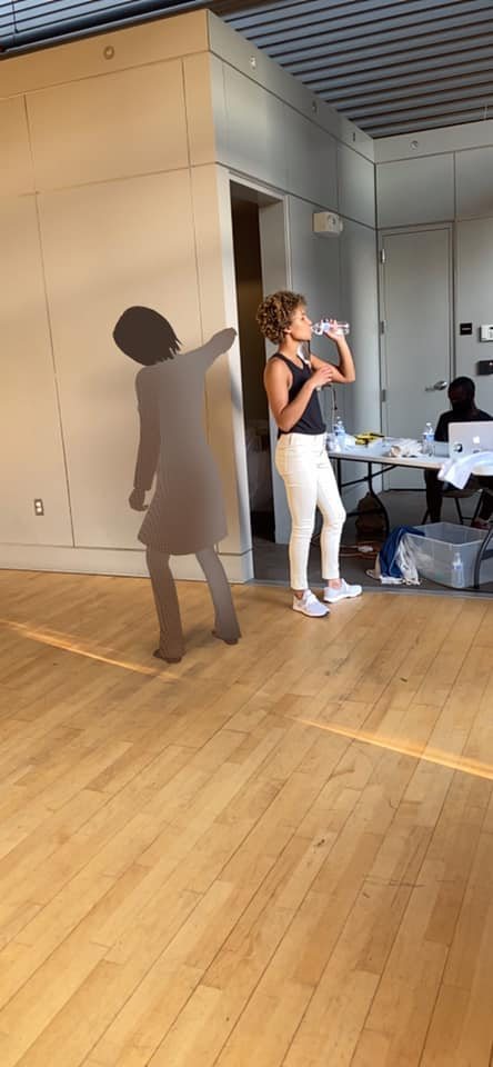 A dancer pausing for water beside an augmented-reality figure on the studio wall