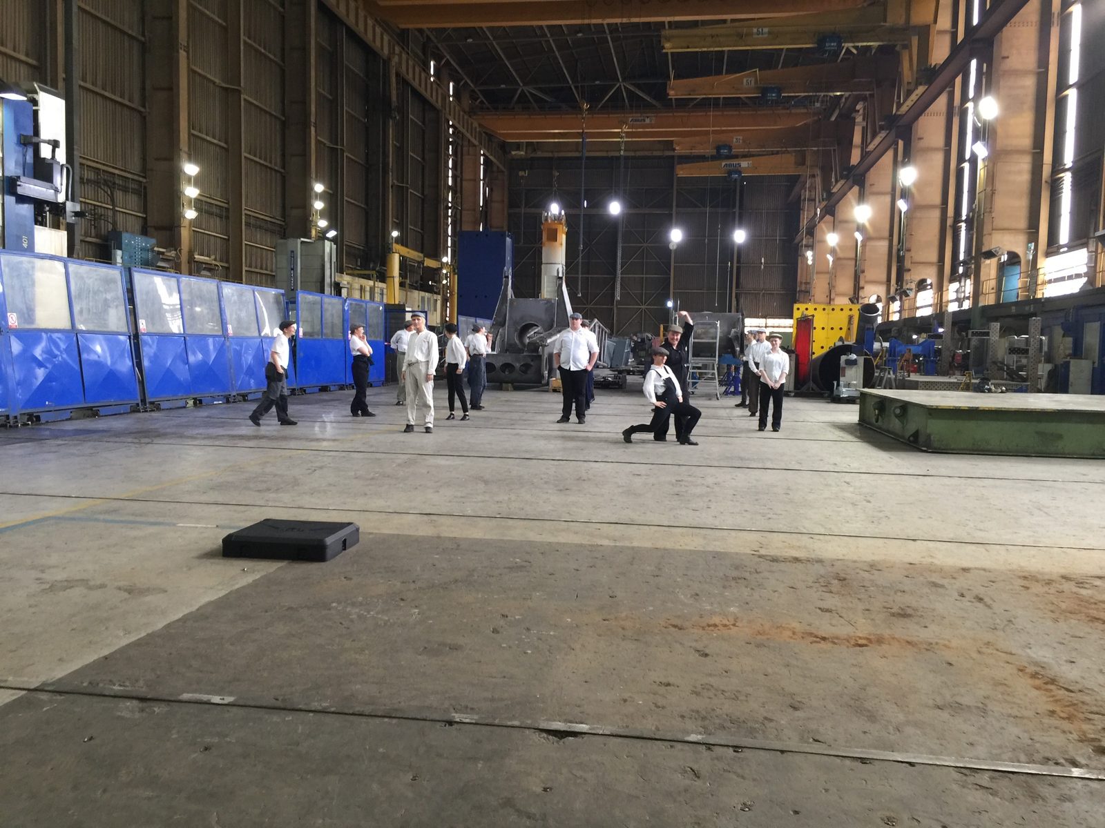 Performers in white shirts and flat caps in the shipyard, one kneeling in a lunge